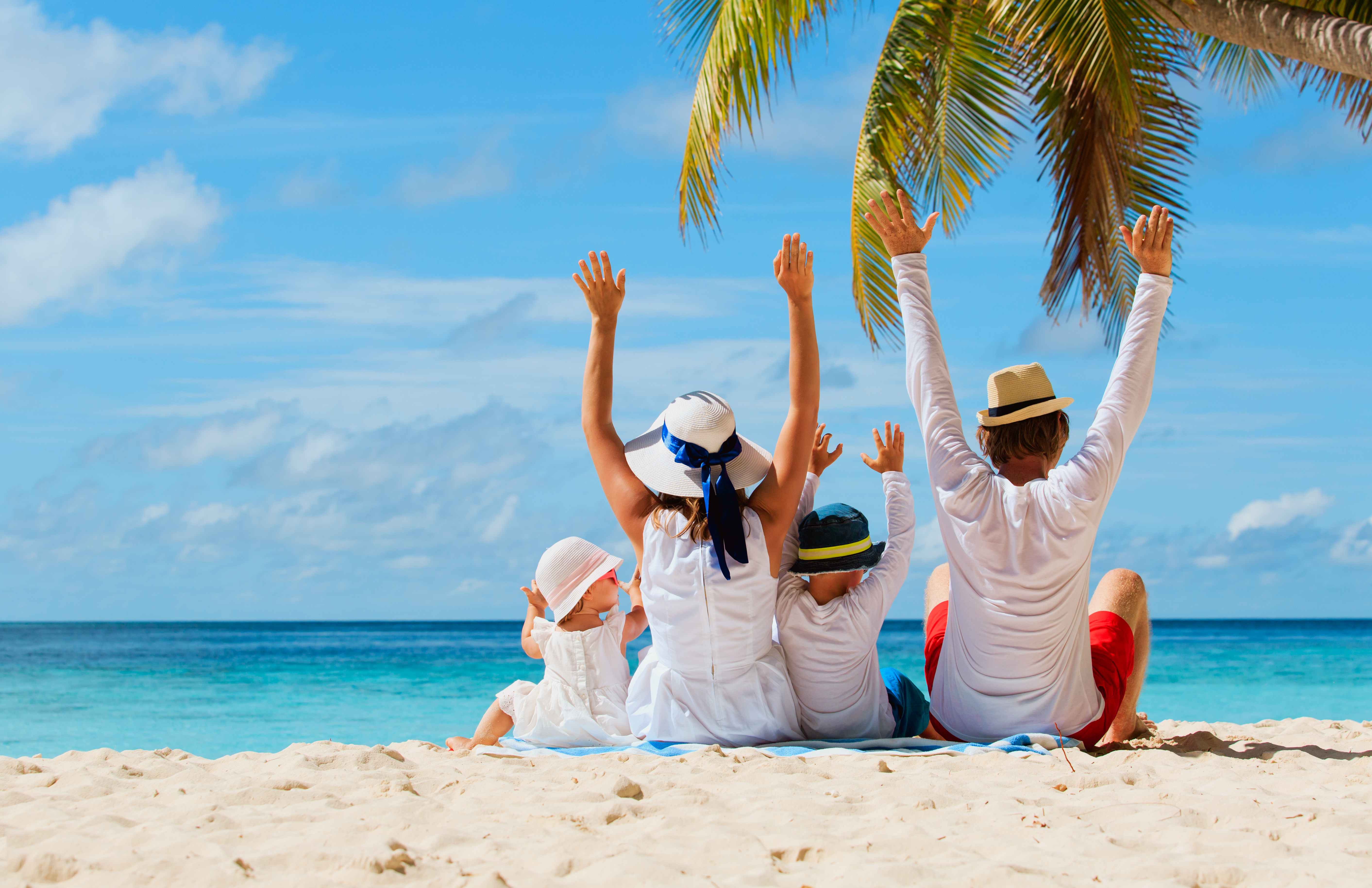 Una famiglia seduta di spalle in spiaggia guarda il mare, le mani alzate al cielo, sereni come quando si fanno viaggi sicuri.